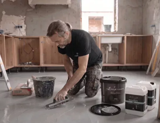 Aplicador profesional extiende microcemento Betoncire BetonBase MN sobre suelo con llana, junto a imprimación BetonPrim 100 durante reforma de cocina.